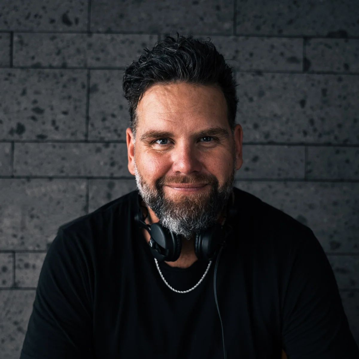 A portrait of Sean Strecker wearing headphones around his neck, smiling against a dark brick wall.